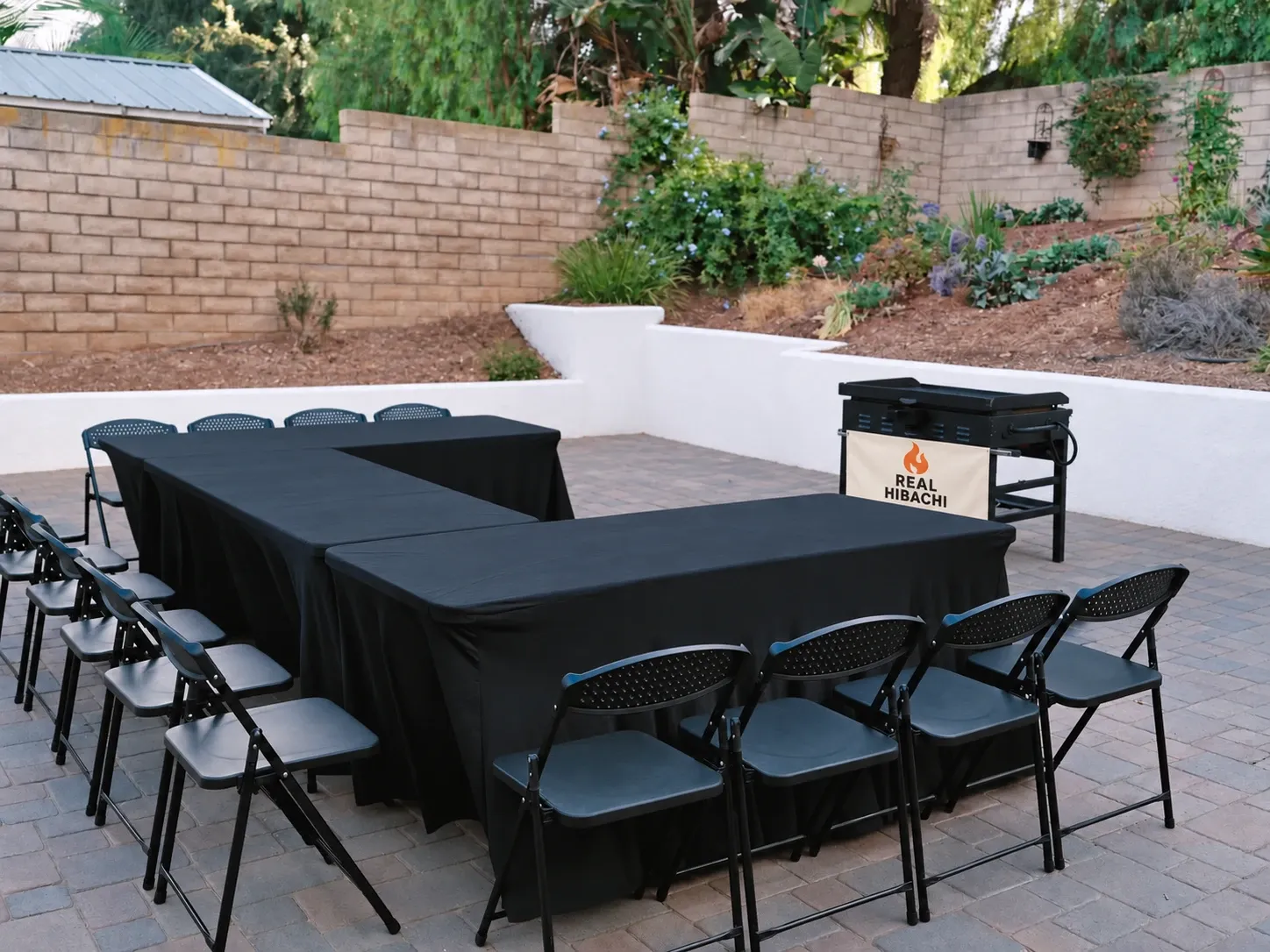 Folding tables in a U shape under black fitted tablecloths with black folding chairs, set up on a backyard patio beside the Real Hibachi grill